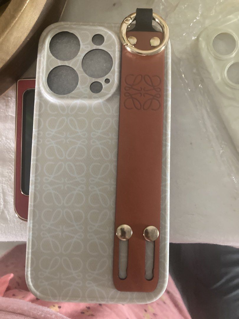 Loewe iPhone Case with Brown Leather Strap (Category: Cases & Sleeves) located in Singapore. Submitted by happysoul9 (Listing ID 1458769694, Image 3). Description: - Loewe phone case set - Suitable for iPhone - Features brown leather strap with gold hardware - Case has a subtle all-over pattern - Includes a clear phone case with a strap - Offers stylish phone protection.